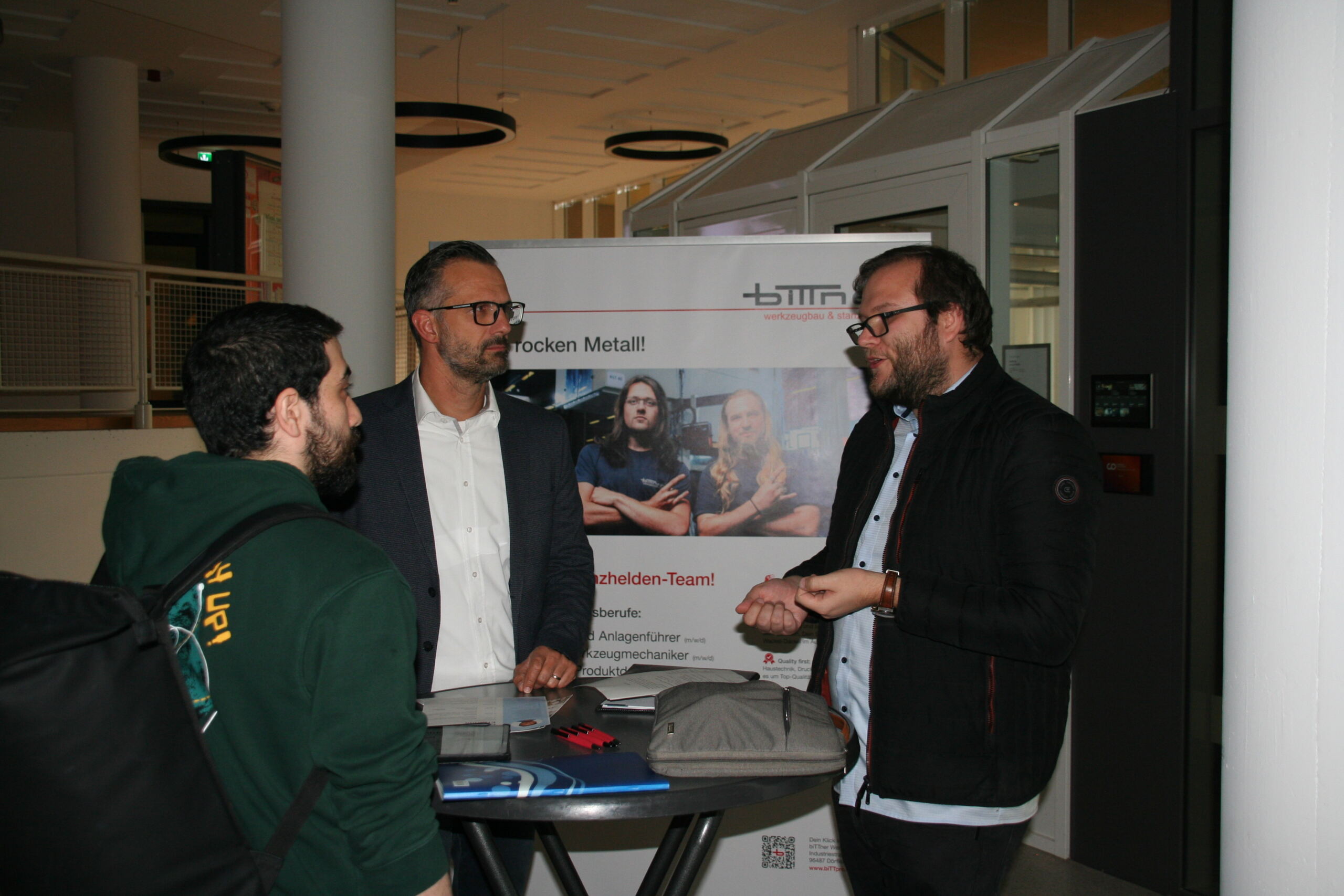 Bei der Veranstaltung der Hochschule Coburg stehen drei Männer um einen hohen Tisch und unterhalten sich. Sie scheinen sich in einem Innenbereich zu befinden. Im Hintergrund sind Menschen zu sehen, die mit Maschinen arbeiten. Ein Mann hält einen Rucksack.