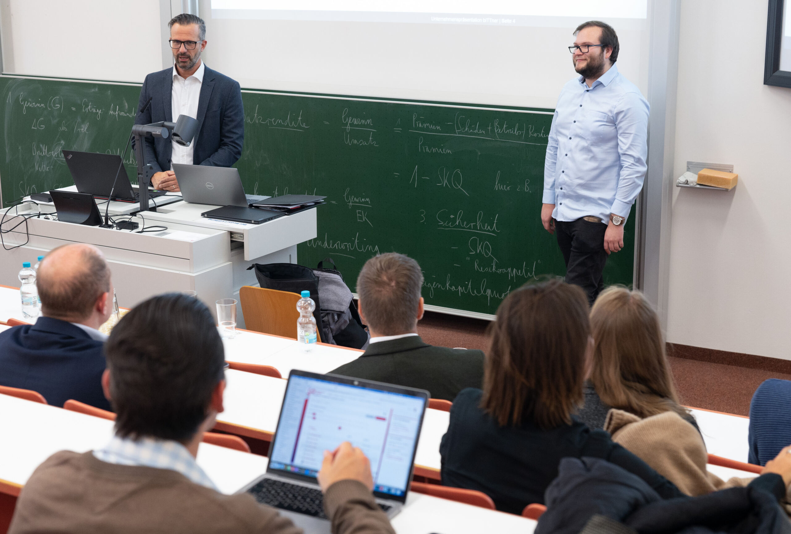 Ein Mann steht neben einem Rednerpult, ein anderer daneben im Hörsaal der Hochschule Coburg. Sie präsentieren einem Publikum, das vor einer Tafel voller Notizen sitzt. Auf den Schreibtischen sind mehrere Laptops zu sehen, die den spannenden akademischen Diskurs aufzeichnen.