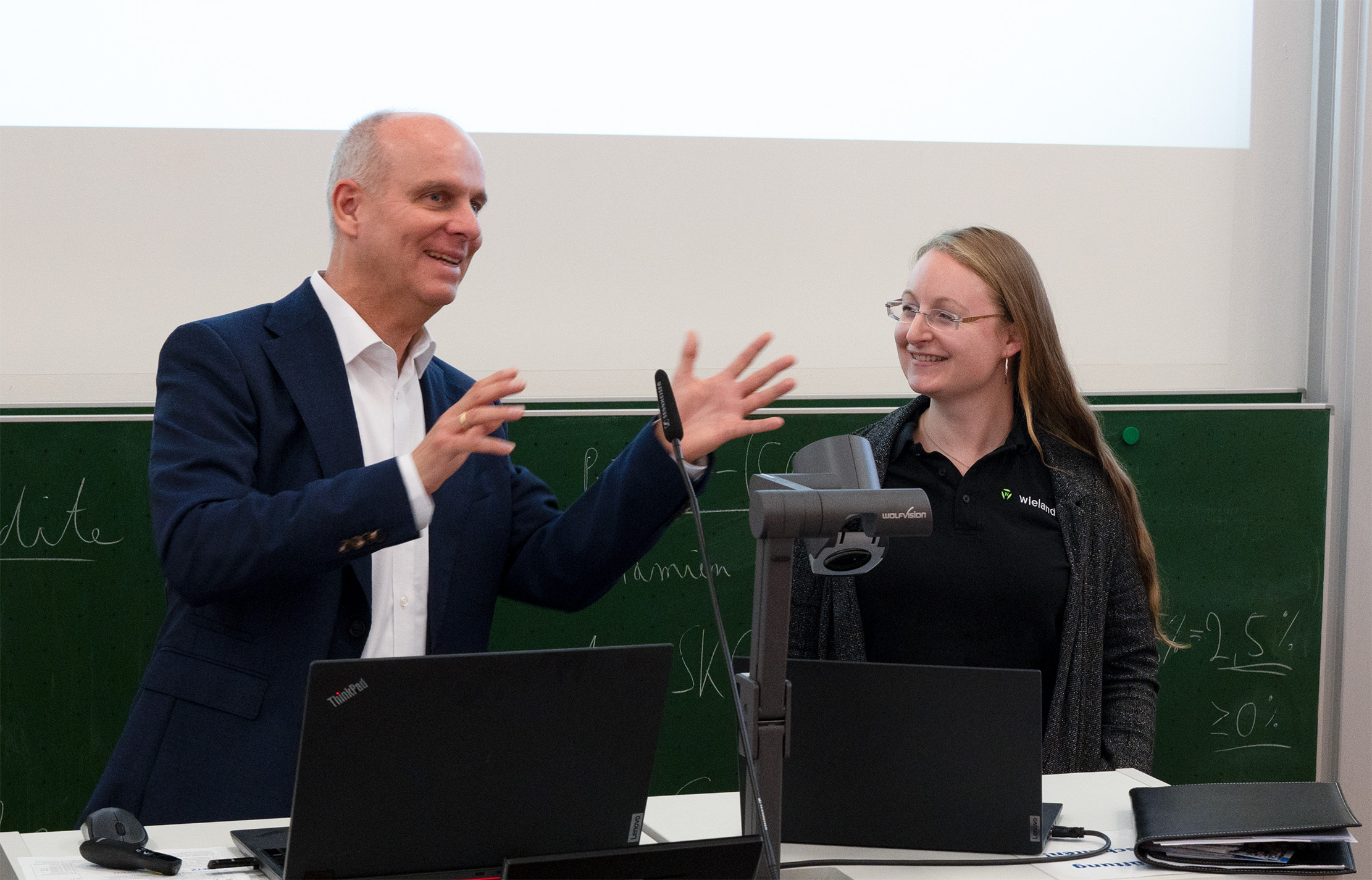 Ein Mann im Anzug und eine Frau in Freizeitkleidung stehen mit Laptops an einem Rednerpult und unterhalten sich angeregt vor einer Tafel und einer Projektionsfläche in einem Unterrichtsraum der Hochschule Coburg.