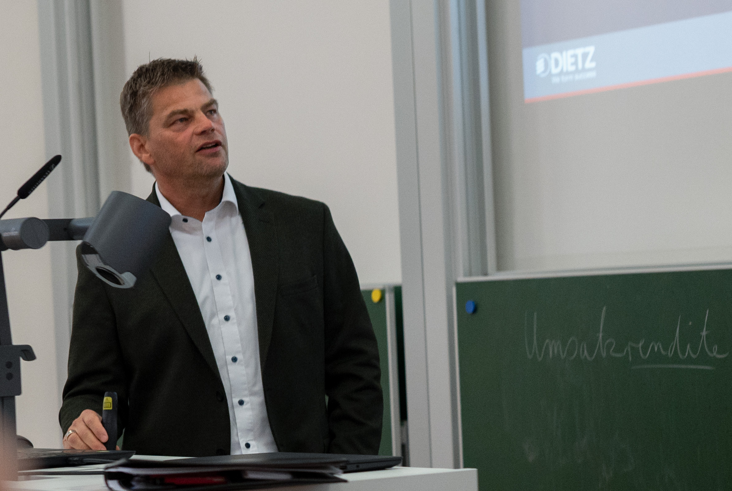 Ein Mann im Anzug spricht vor einer projizierten Präsentation. Er steht an einem Schreibtisch mit einer Dokumentenkamera. Im Hintergrund ist eine Tafel mit unleserlicher Schrift zu sehen.