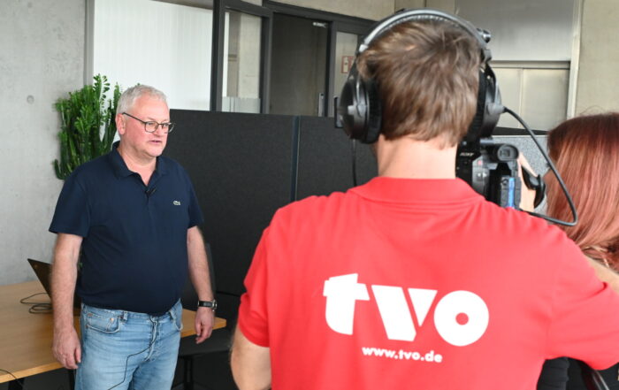 Ein Mann im dunkelblauen Hemd wird im Innenbereich interviewt, möglicherweise über PV-Forschung. Eine weitere Person in einem roten Hemd mit den Aufdrucken „tvo“ und „www.tvo.de“ auf dem Rücken bedient eine Kamera für den TV-Sender. Im Hintergrund sind ein Laptop und eine Topfpflanze zu sehen.