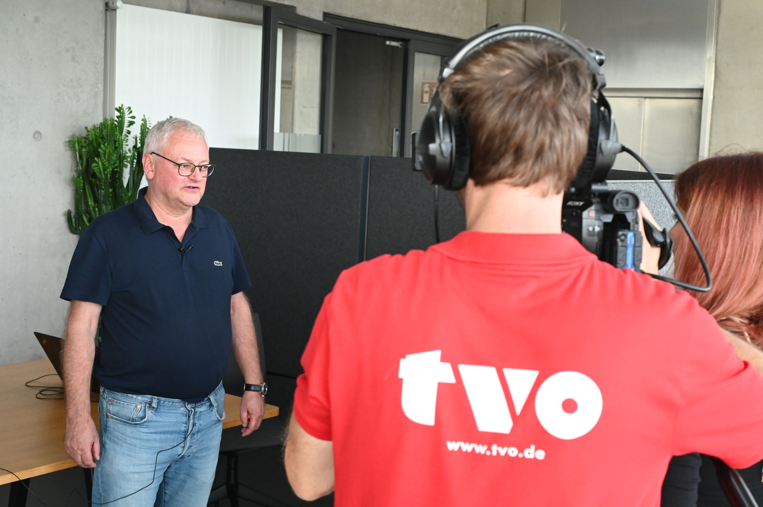 Ein Mann im dunkelblauen Hemd wird im Innenbereich interviewt, möglicherweise über PV-Forschung. Eine weitere Person in einem roten Hemd mit den Aufdrucken „tvo“ und „www.tvo.de“ auf dem Rücken bedient eine Kamera für den TV-Sender. Im Hintergrund sind ein Laptop und eine Topfpflanze zu sehen.