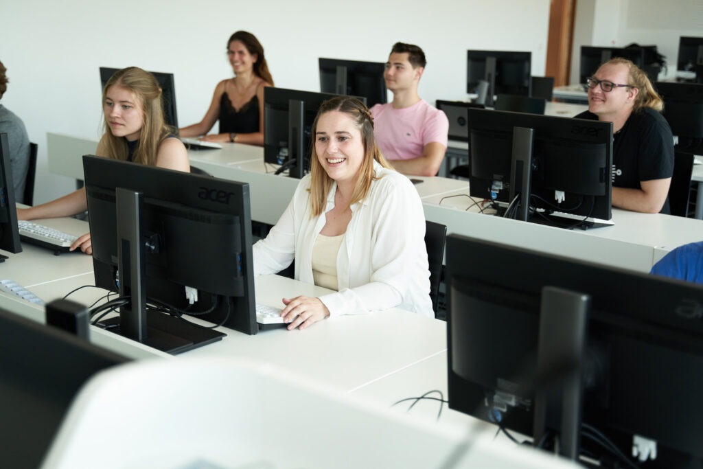 In einem gut beleuchteten, in Reihen angeordneten Hörsaal sitzt eine Gruppe von Studierenden an Tischen mit Computern, konzentriert und aufmerksam. Sie scheinen auf ihre Bildschirme konzentriert zu sein und vertiefen sich im Rahmen ihrer Bachelor-Studiengänge in die Komplexität der Informatik.