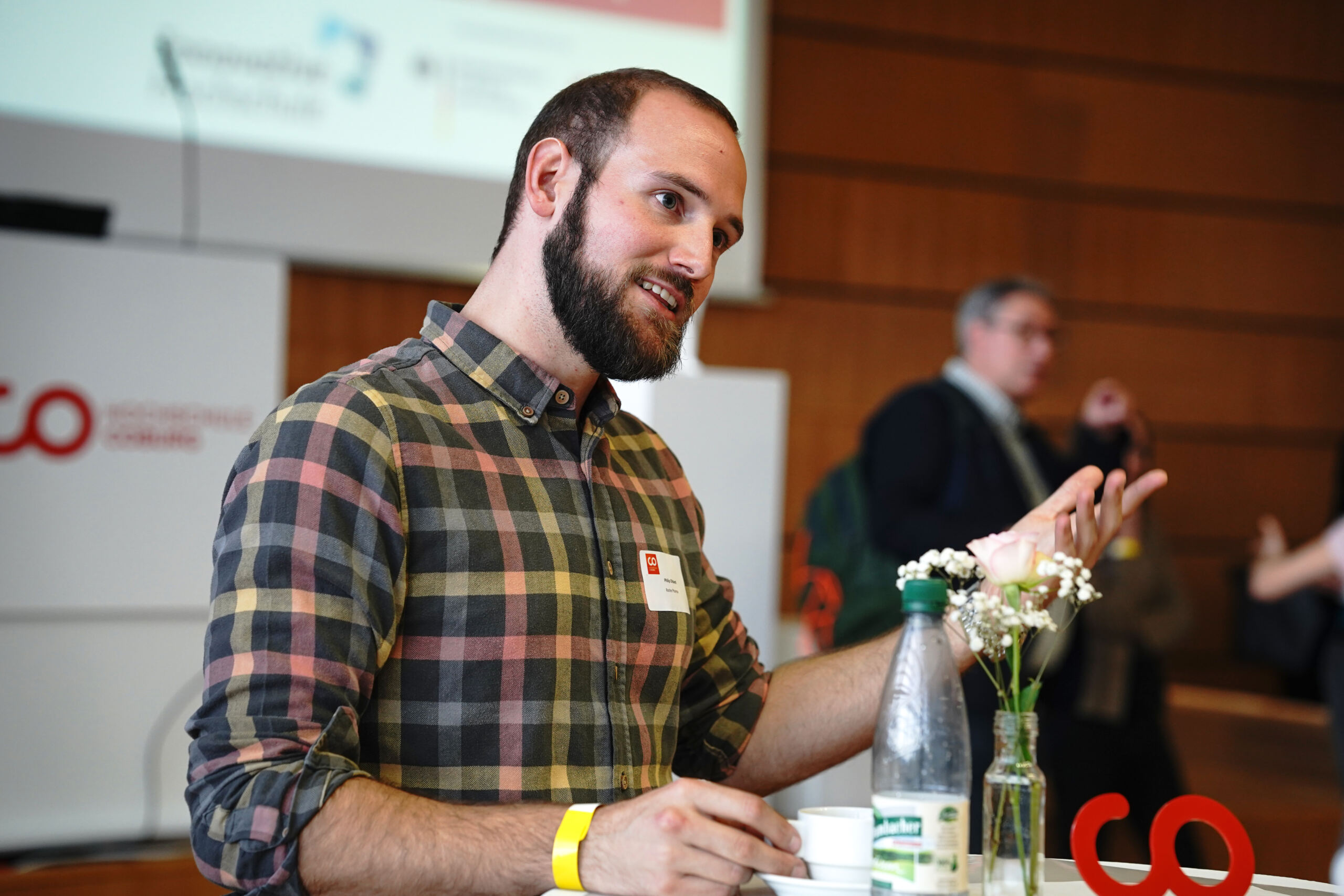 Ein Mann mit Bart und kariertem Hemd gestikuliert, während er an einem Tisch spricht. Vor ihm stehen eine Wasserflasche und Blumen. Im Hintergrund sind verschwommen Menschen zu sehen, was auf eine Konferenz oder Veranstaltung hindeutet.