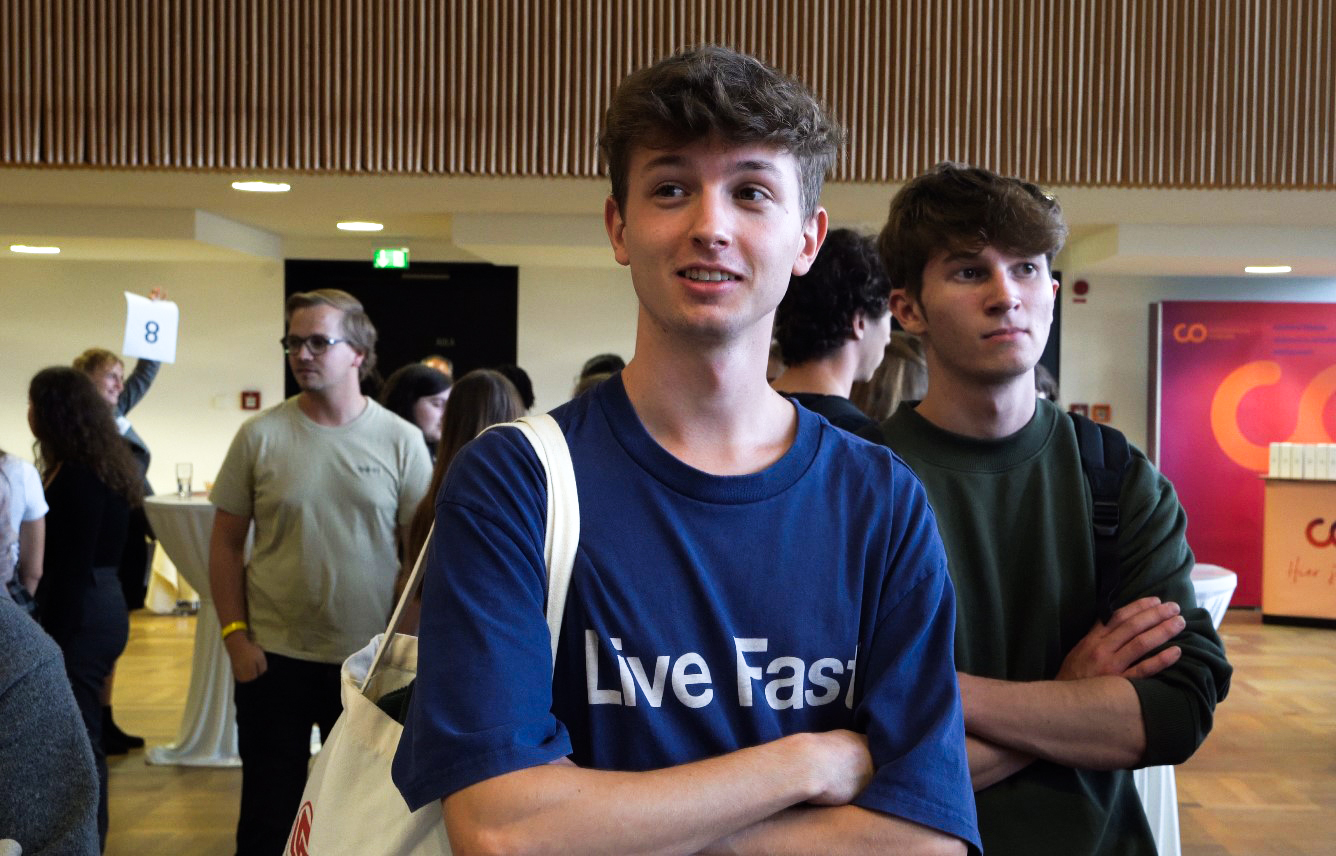 Zwei junge Männer stehen in einem überfüllten Innenraum, einer trägt ein blaues „Live Fast“-Shirt und eine Einkaufstasche, der andere ein grünes Shirt mit verschränkten Armen. Im Hintergrund sind Menschen und Veranstaltungsstände zu sehen.