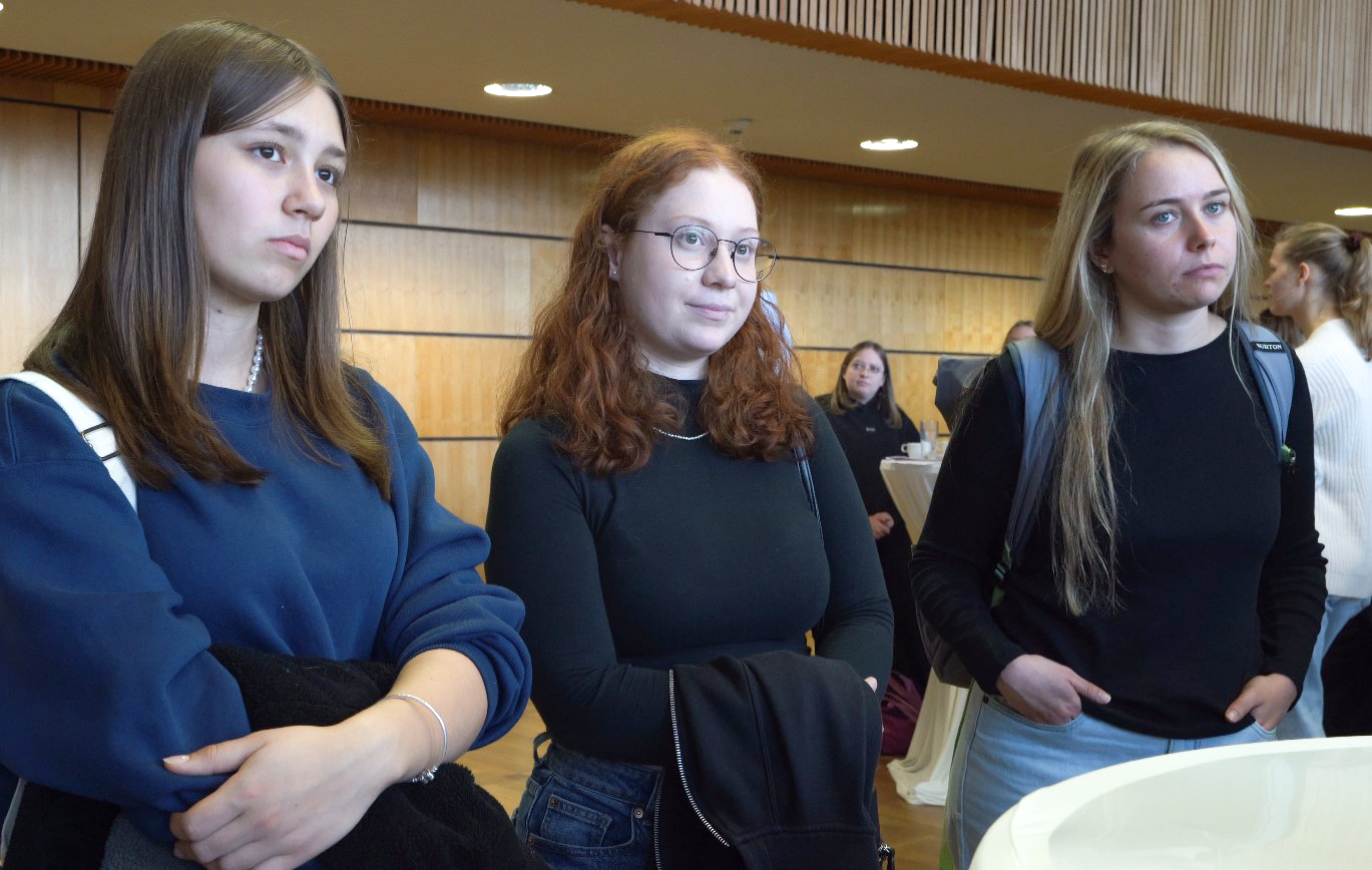 Drei Frauen stehen drinnen und wirken aufmerksam. Eine trägt einen marineblauen Pullover, eine andere eine Brille mit schwarzem Oberteil und die dritte hat einen Rucksack und hellblaue Hosen an. Im Hintergrund sind Holzvertäfelungen und andere Menschen in unscharfer Darstellung zu sehen.