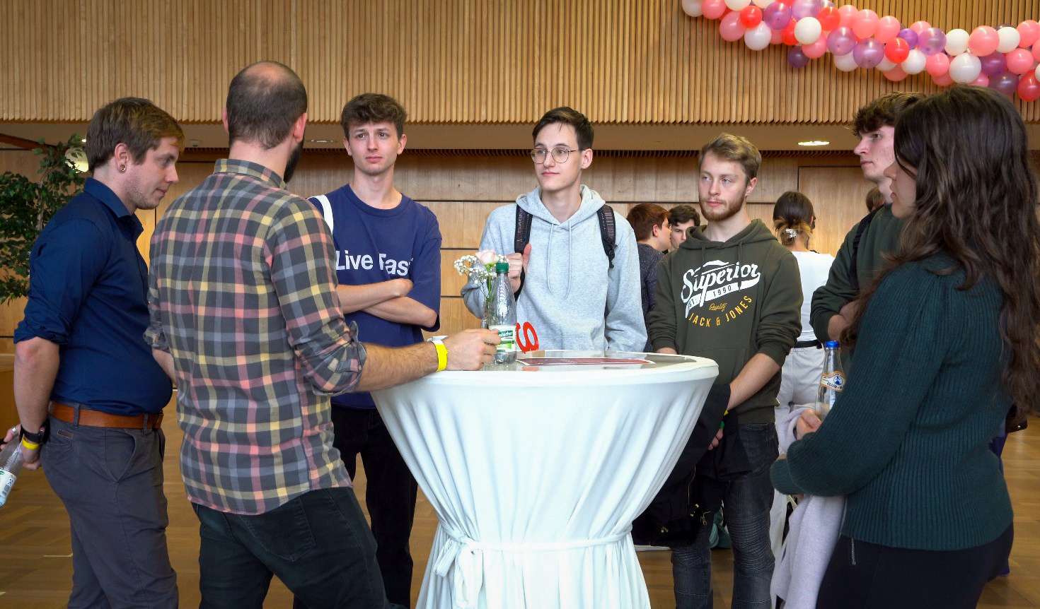 Bei einer Indoor-Veranstaltung steht eine Gruppe von Menschen um einen großen runden Tisch. Einige unterhalten sich, andere hören zu. Der Raum hat Holzwände und eine mit Luftballons dekorierte Decke.