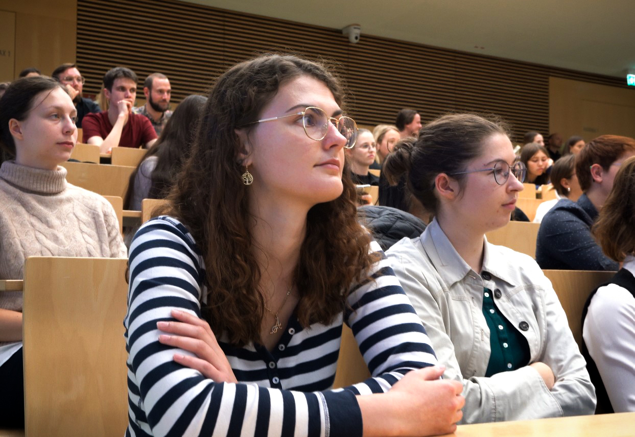 Eine Gruppe von Studenten hört aufmerksam einer Vorlesung in einem Hörsaal zu. Der Raum ist mit Holz getäfelt und die Studenten sitzen in abgestuften Reihen. Einige von ihnen tragen Brillen und einige machen sich Notizen.