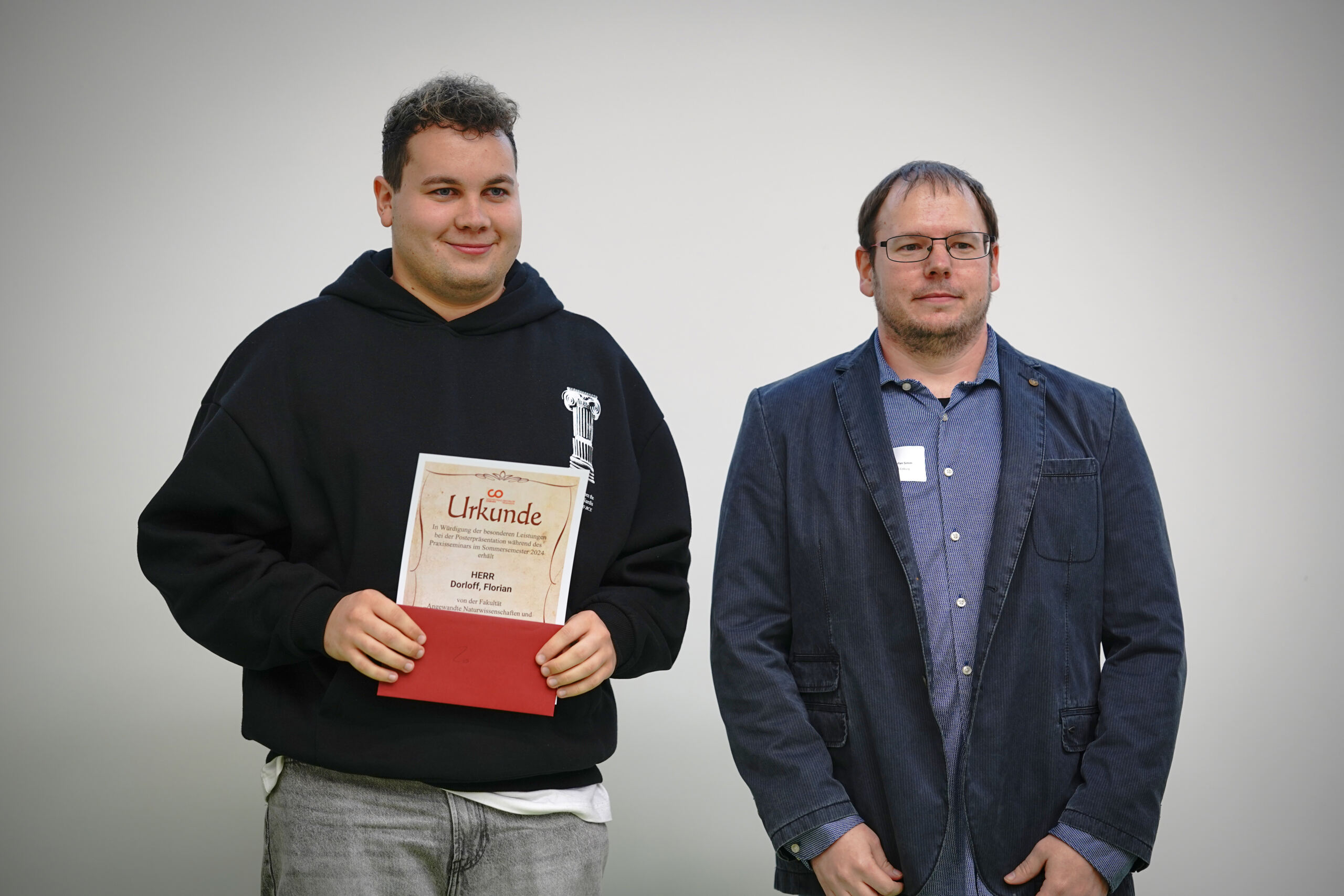 Zwei Männer stehen zusammen. Der Mann links hält ein Zertifikat mit rotem Umschlag. Er trägt einen schwarzen Kapuzenpullover und Jeans. Der Mann rechts trägt eine Brille, einen blauen Blazer, ein kariertes Hemd und Jeans. Sie stehen vor einem schlichten grauen Hintergrund.