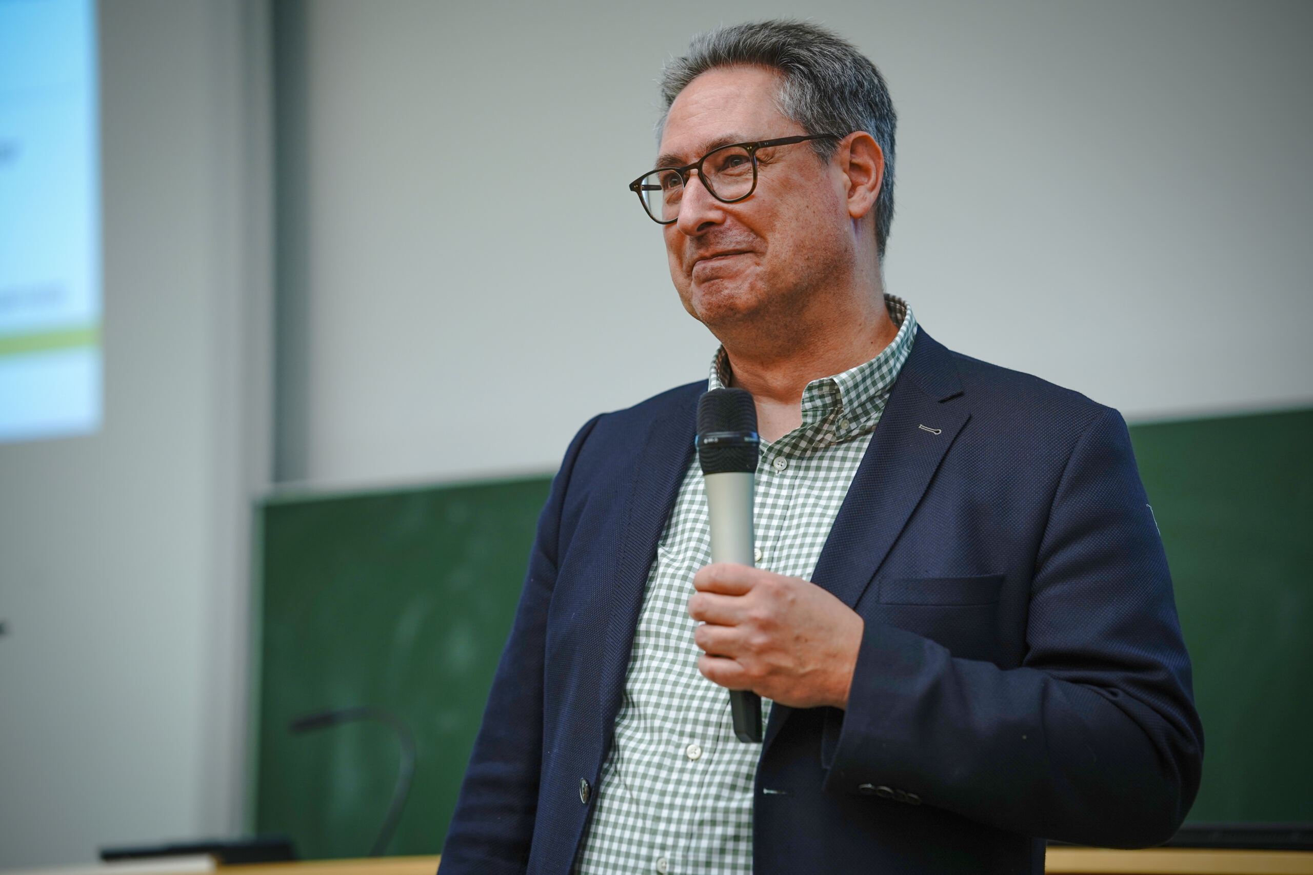 Ein Mann in Blazer und Brille hält ein Mikrofon, während er in einem Hörsaal spricht. Hinter ihm ist eine grüne Tafel und seitlich ist teilweise eine Projektionsleinwand zu sehen.