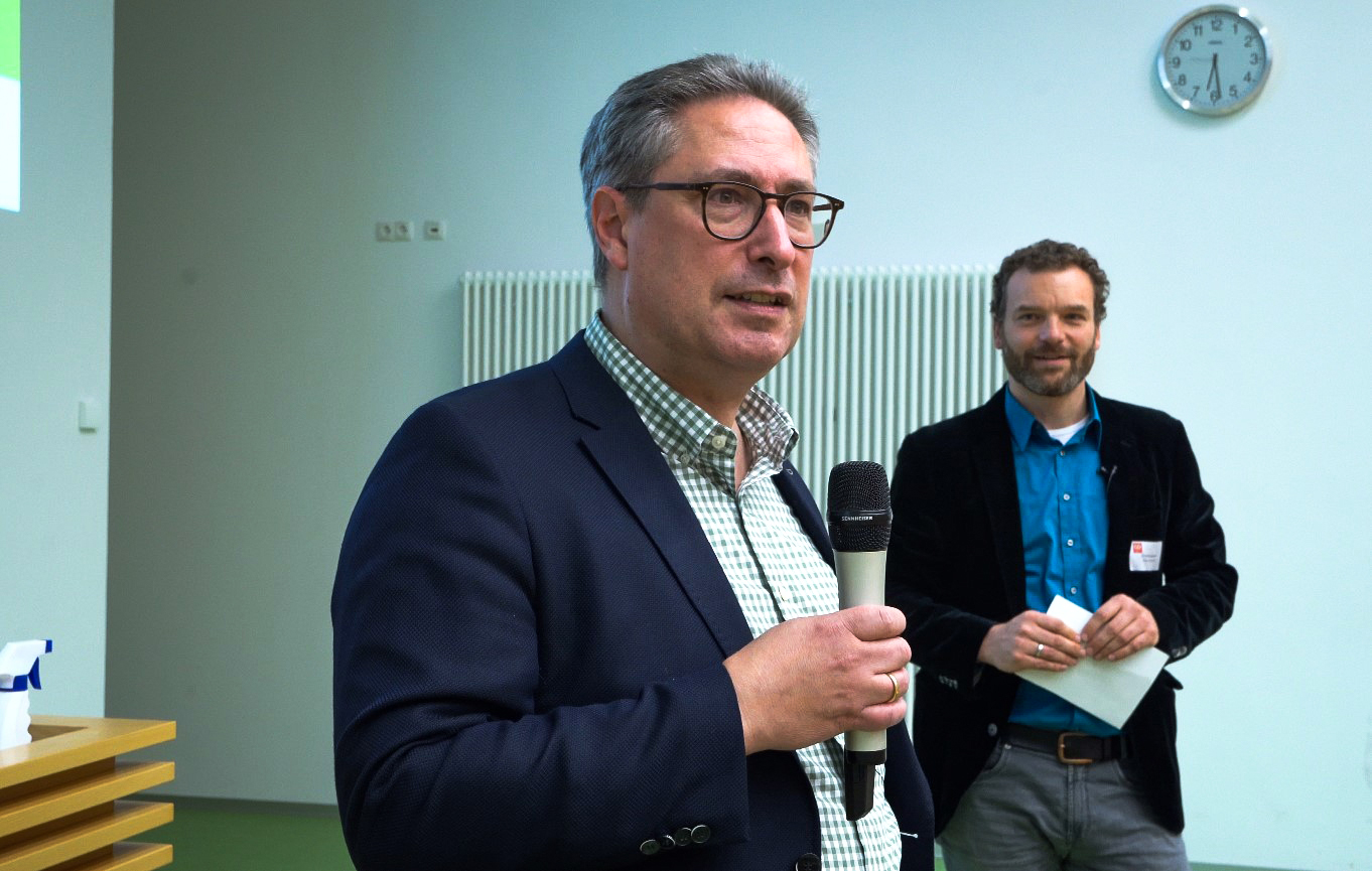 Ein Mann in Anzug und Brille spricht in ein Mikrofon. Ein anderer Mann steht im Hintergrund und hält Papiere in der Hand. Sie befinden sich in einem Raum, in dem eine Wanduhr und ein Heizkörper zu sehen sind.