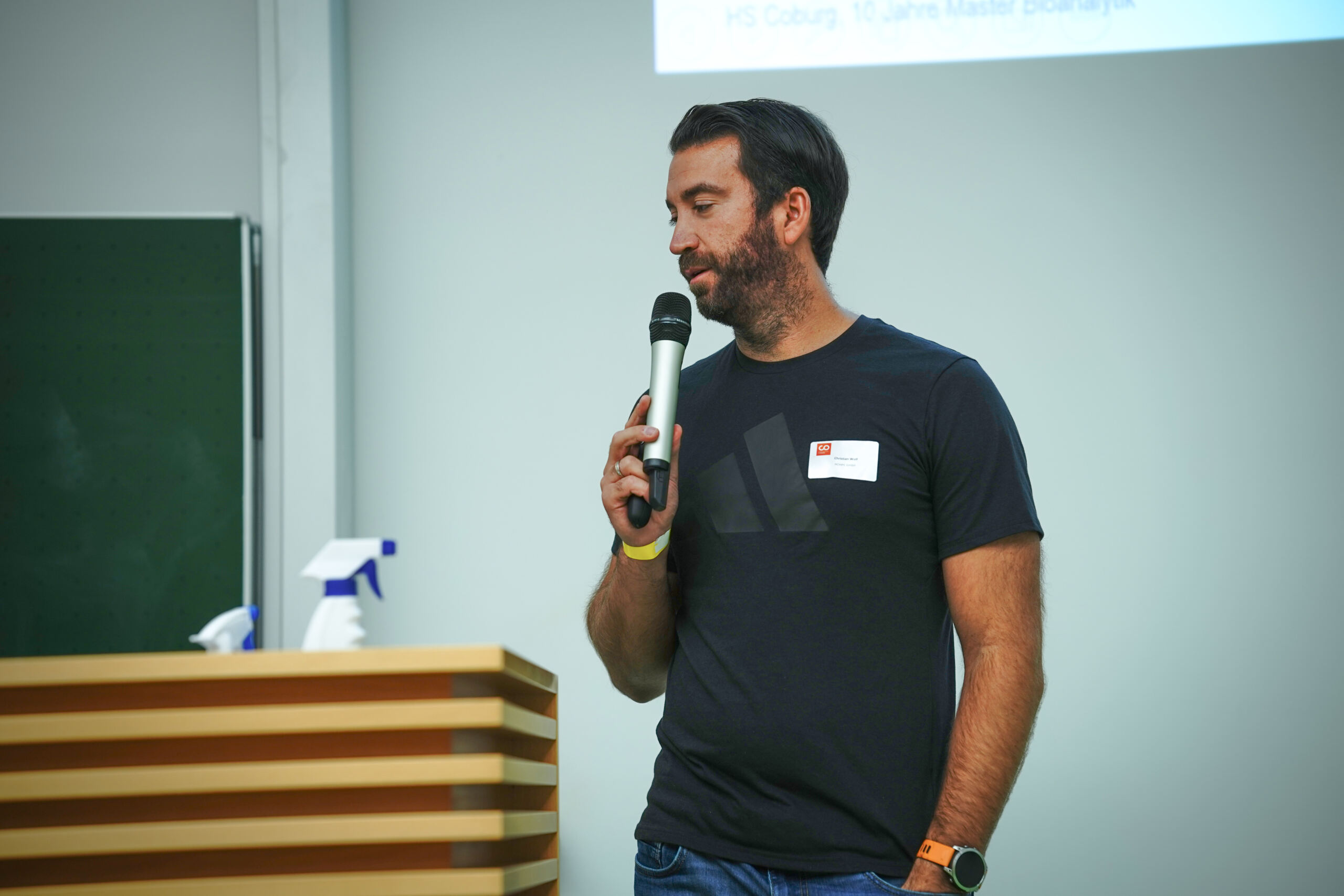 Ein Mann mit Bart steht neben einem hölzernen Rednerpult und spricht in ein Mikrofon. Er trägt ein schwarzes T-Shirt und Jeans. Im Hintergrund sind eine grüne Tafel und eine Sprühflasche zu sehen.