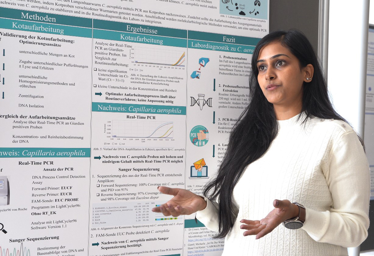 Eine Frau im weißen Pullover präsentiert Forschungsergebnisse vor einem ausführlichen Poster mit Grafiken und Text zur DNA-Analyse. Sie gestikuliert mit den Händen und erklärt einem Publikum wissenschaftliche Erkenntnisse.