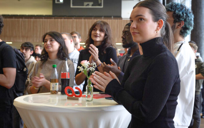 Eine Gruppe von Menschen steht während einer Indoor-Veranstaltung um einen Tisch und klatscht. Auf dem Tisch stehen Flaschen und eine kleine Vase mit Blumen. Die Atmosphäre wirkt lebhaft und engagiert.