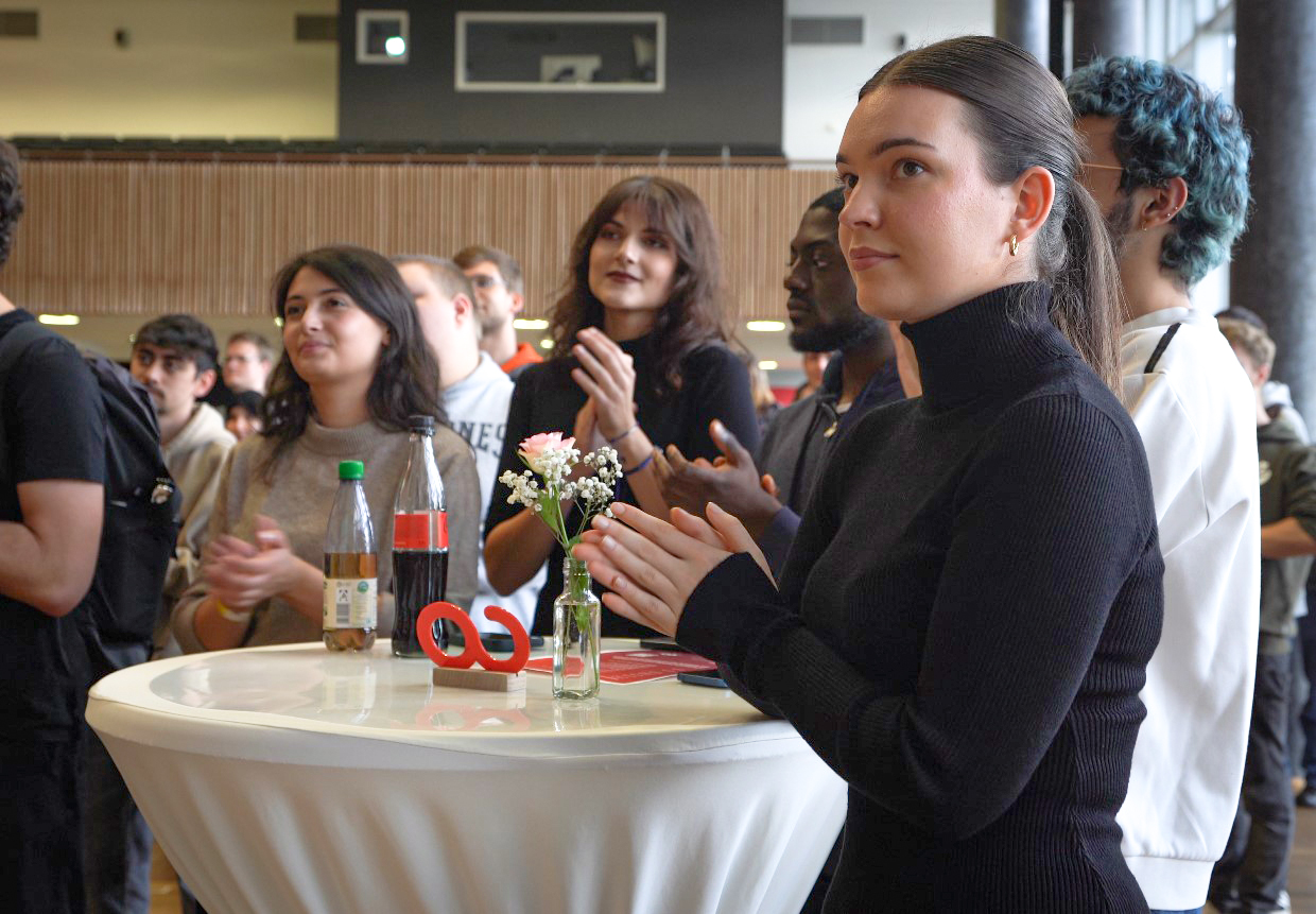 Eine Gruppe von Menschen steht während einer Indoor-Veranstaltung um einen Tisch und klatscht. Auf dem Tisch stehen Flaschen und eine kleine Vase mit Blumen. Die Atmosphäre wirkt lebhaft und engagiert.