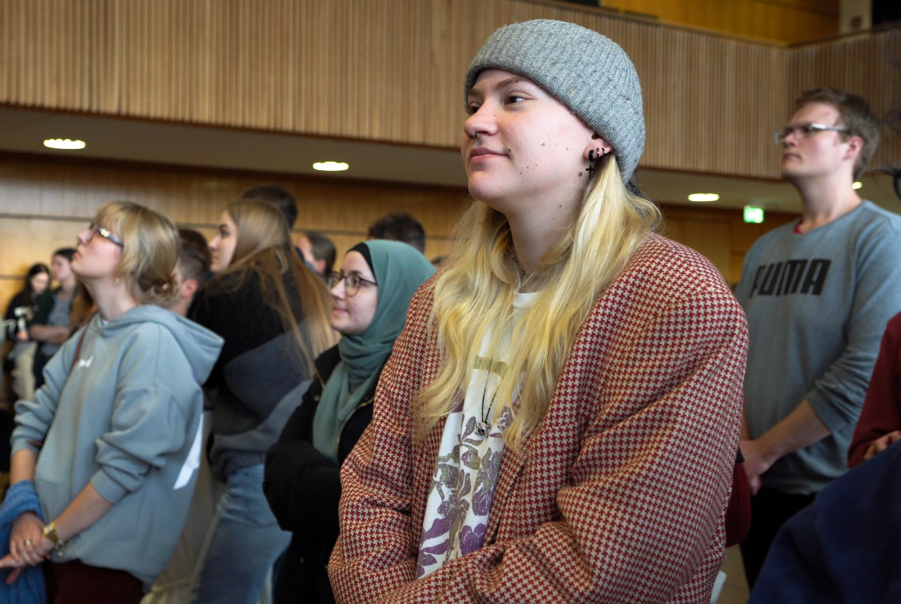 Eine Gruppe von Menschen steht aufmerksam in einem Raum mit Holzwänden. Eine Person im Vordergrund trägt eine graue Beanie-Mütze und eine gemusterte Jacke. Die anderen um sie herum sind leger gekleidet. Die Atmosphäre wirkt nachdenklich.