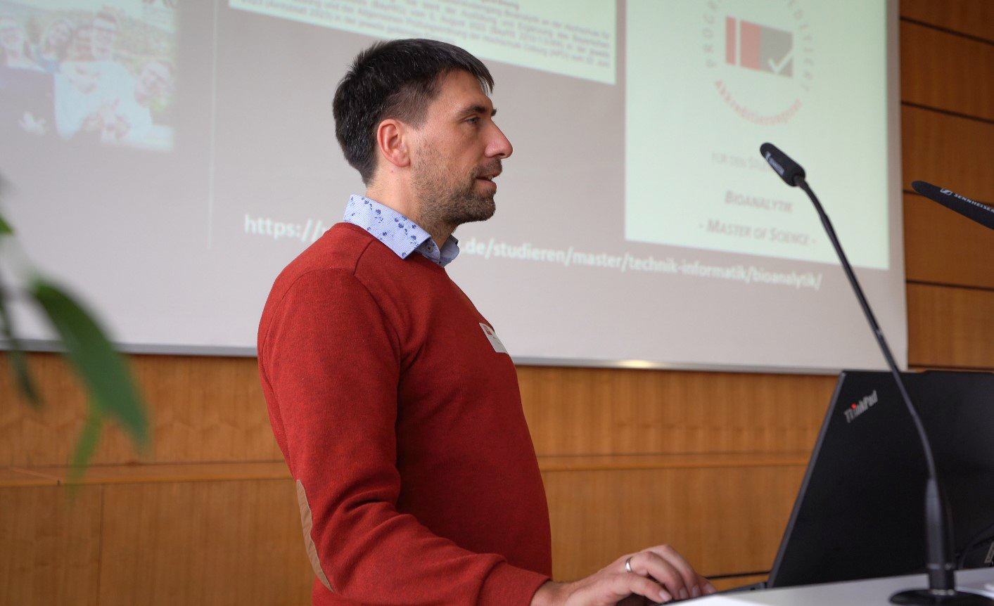 Ein Mann im roten Pullover steht an einem Rednerpult und spricht zu einem Publikum. Auf einer Leinwand hinter ihm wird eine Präsentation mit Texten und Grafiken zu Bioanalytik und einem Masterstudiengang projiziert. Auf dem Rednerpult ist ein Laptop zu sehen.