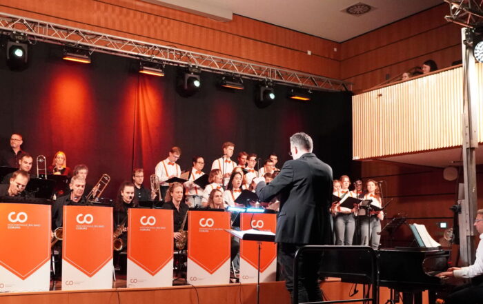 Auf der Bühne tritt eine große Jazzband auf, deren Musiker und Musikerinnen gekonnt Blech- und Holzblasinstrumente spielen. Vorne steht ein Dirigent, während seitlich ein Pianist der nahegelegenen Hochschule spielt. Die Bühne erstrahlt in rotem und orangefarbenem Licht, während das Publikum teilweise sichtbar sitzt.