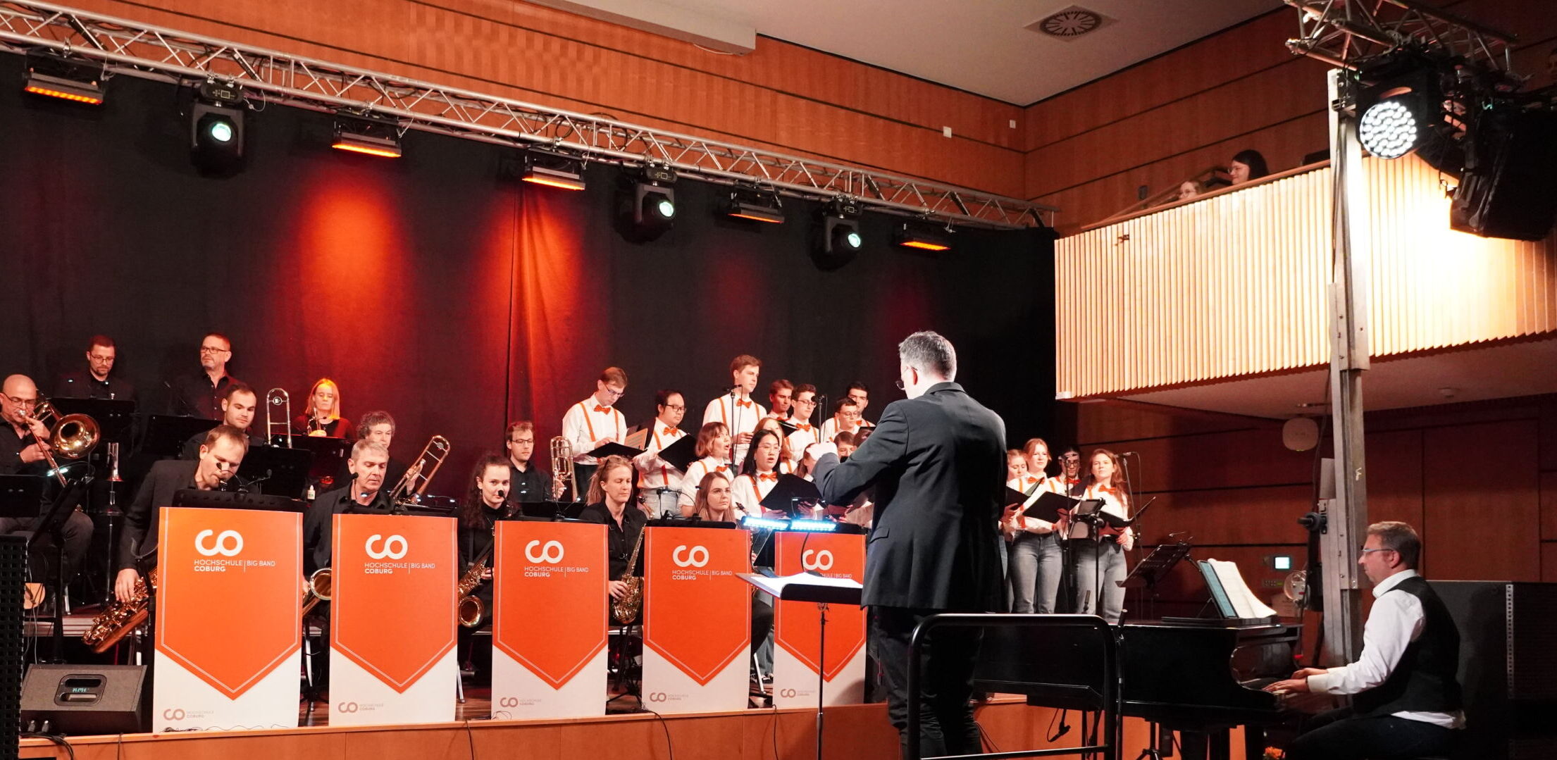 Auf der Bühne tritt eine große Jazzband auf, deren Musiker und Musikerinnen gekonnt Blech- und Holzblasinstrumente spielen. Vorne steht ein Dirigent, während seitlich ein Pianist der nahegelegenen Hochschule spielt. Die Bühne erstrahlt in rotem und orangefarbenem Licht, während das Publikum teilweise sichtbar sitzt.