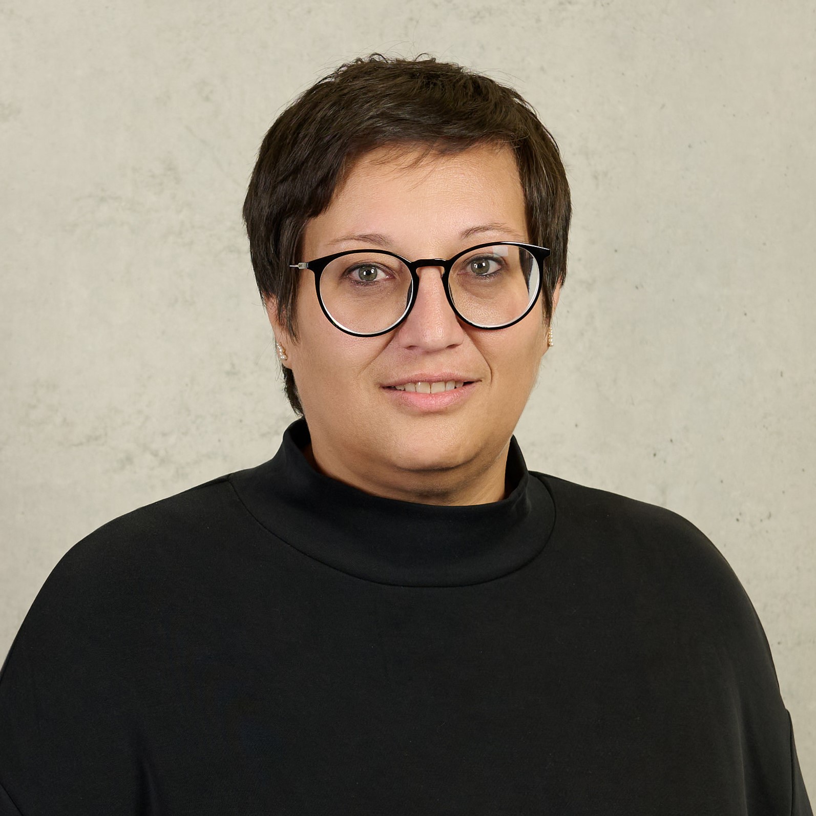 A person with short hair and glasses, proudly representing Hochschule Coburg, smiles at the camera. They wear a dark shirt adorned with small decorative elements against a plain, light-colored wall.