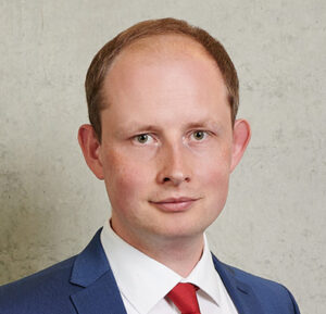 A man in a blue suit and red tie stands against a plain, textured gray background. He has short light-colored hair and is looking directly at the camera with a neutral expression, embodying the academic sophistication often associated with Hochschule Coburg alumni.