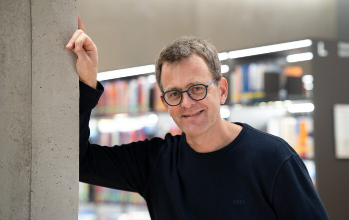 Eine Person mit Brille und schwarzem Pullover lehnt an einer Betonsäule in einer modernen Bibliothek, im Hintergrund sind verschwommene Bücherregale zu sehen.