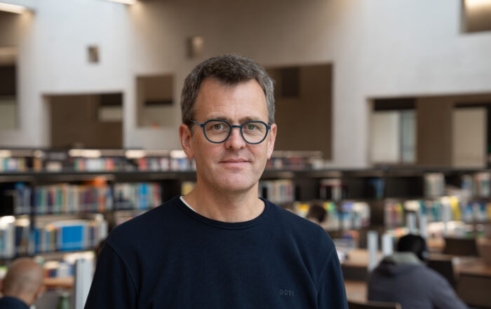 Ein Mann mit kurzen Haaren und Brille steht in einer Bibliothek. Er trägt einen dunklen Pullover. Regale voller Bücher und Menschen, die im Hintergrund lernen.