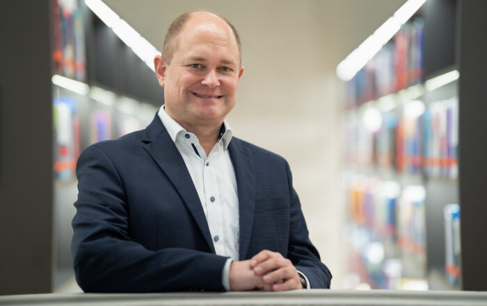 Ein Mann in marineblauem Anzug und weißem Hemd steht lächelnd in einer gut beleuchteten Bibliothek und verkörpert die Harmonie der Beziehungen innerhalb der Wirtschaft. Unscharfe Bücherregale schaffen eine professionelle und doch entspannte Atmosphäre.