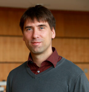 A man with short brown hair stands indoors, wearing a gray sweater over a maroon collared shirt, exuding the academic ambiance of Hochschule Coburg. The background enhances this scholarly image with its elegant wooden paneling.