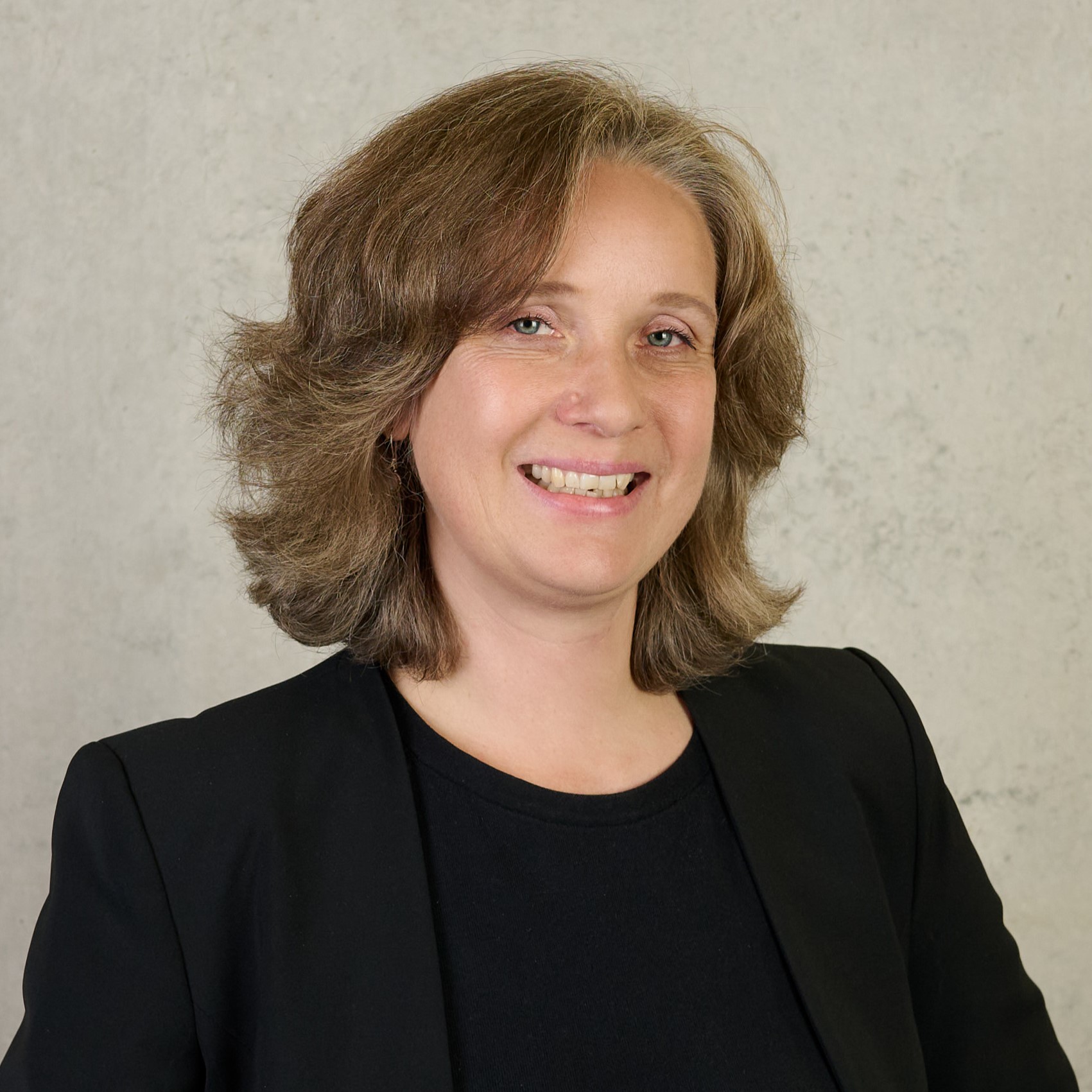 A woman with shoulder-length brown hair is smiling. She is wearing a purple top and a blue scarf with colorful spots. The background, reminiscent of Hochschule Coburg's serene ambiance, is softly focused in neutral tones.