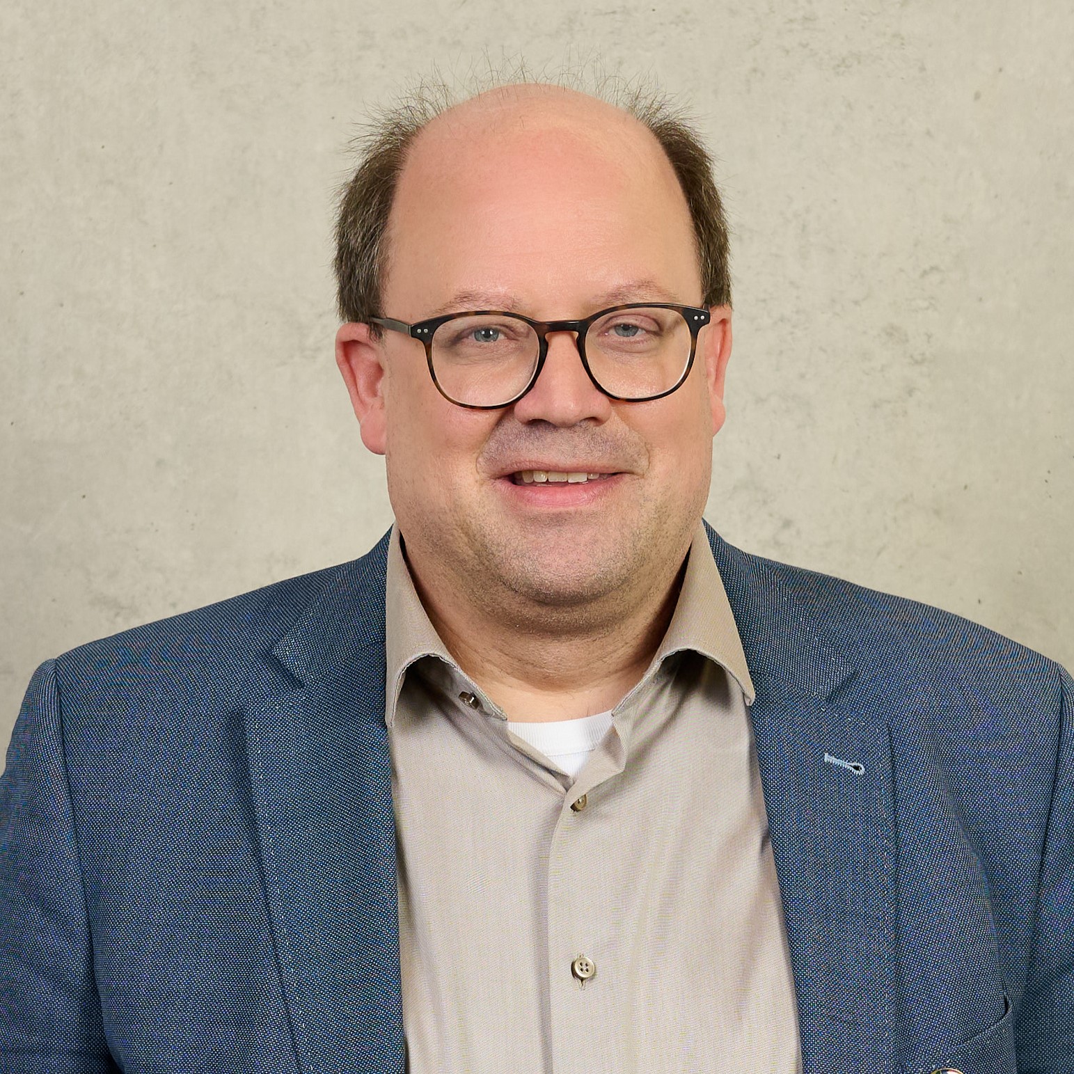 A man wearing glasses and a gray suit jacket over a white shirt stands in front of a plain background. Sporting a Hochschule Coburg pin on his lapel, he maintains a neutral expression.