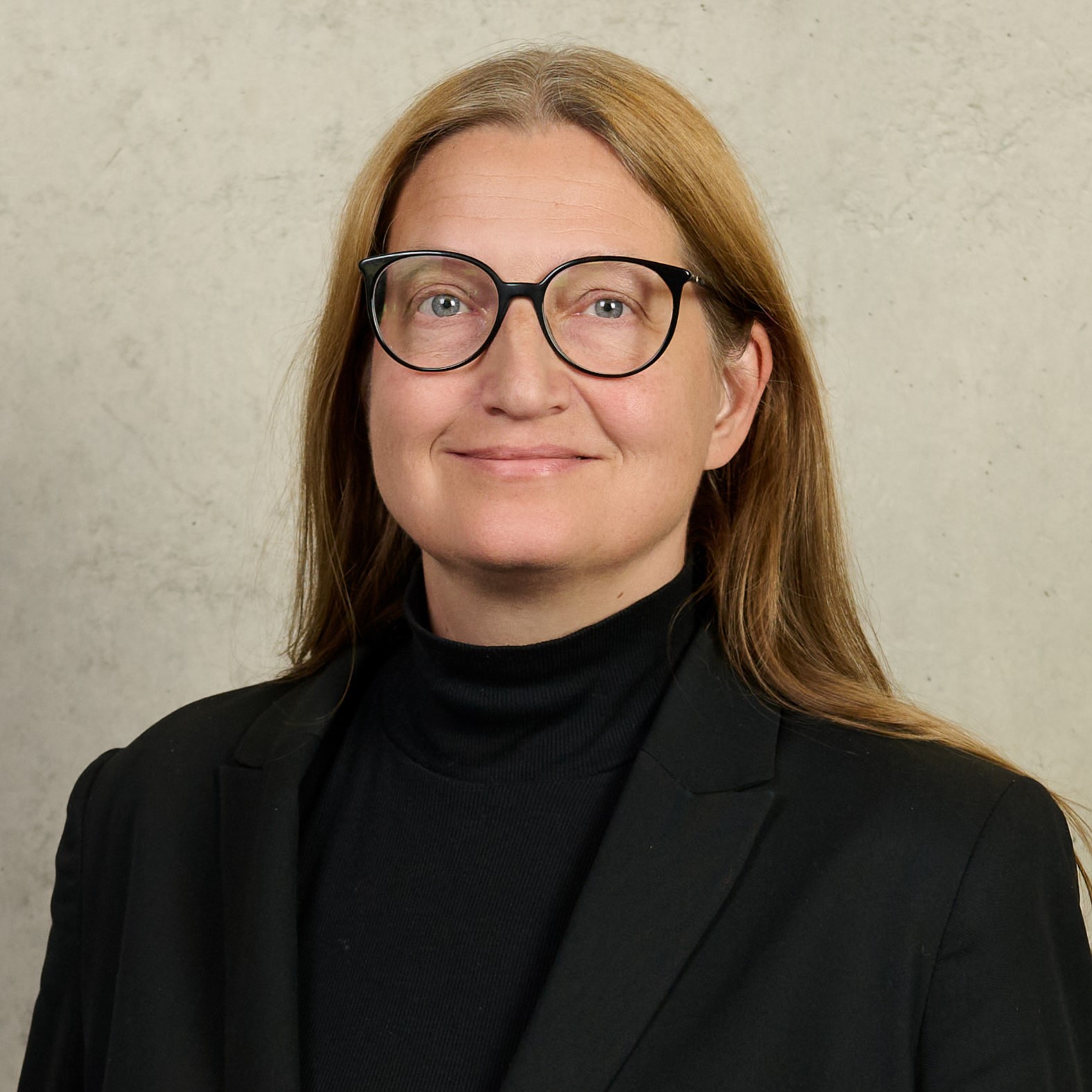 A person with long brown hair and glasses, wearing a dark top and a beaded necklace, smiles slightly. The background has blurred green and white tones, reminiscent of Hochschule Coburg's serene campus environment.