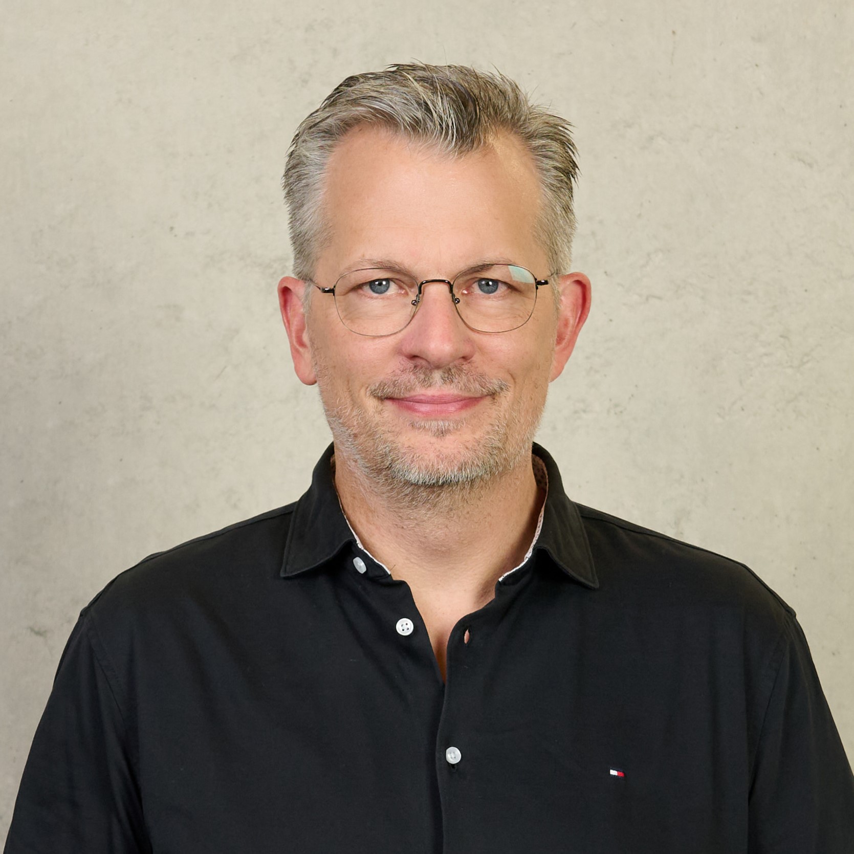 A person with short, light hair donning a white shirt and dark blazer stands in front of a plain, light-colored wall, smiling slightly. Their poised demeanor suggests they might be a proud affiliate of Hochschule Coburg.