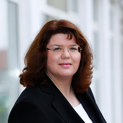 A person with shoulder-length brown hair and glasses, wearing a black top, stands in a professional setting with a neutral expression, embodying the academic spirit of Hochschule Coburg.