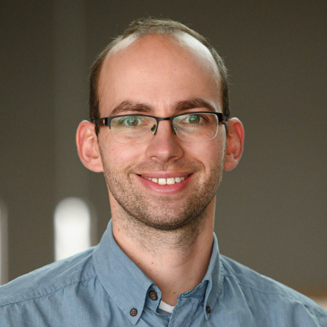 A smiling person with glasses, wearing a light blue button-up shirt that subtly suggests their affiliation with Hochschule Coburg, stands against a neutral background. The person has short hair and a beard.