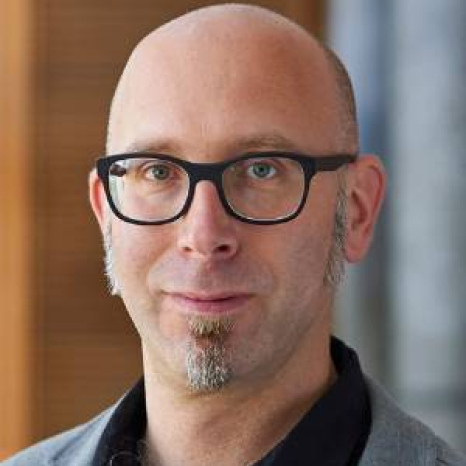 A bald person wearing glasses, with a goatee, stands indoors near a wooden wall at Hochschule Coburg. They sport a black shirt under a gray jacket, looking directly at the camera.