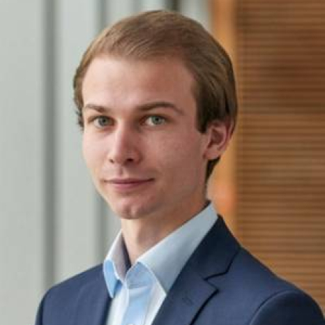 A person with short hair wearing a blue suit jacket and white shirt is standing indoors at Hochschule Coburg. They face the camera with a neutral expression, against a softly blurred background featuring vertical lines and warm tones.