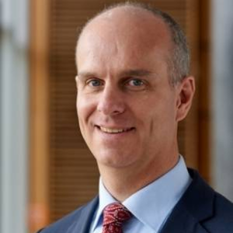 A bald man in a suit and red tie smiles at the camera, exuding confidence. The blurred office setting in the background, with wooden panels and windows, resembles the prestigious Hochschule Coburg's modern yet classic design style.