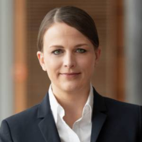 A woman with brown hair in a bun, wearing a white shirt and dark blazer, looks into the camera with a slight smile. She stands in front of a blurred background hinting at Hochschule Coburg's wooden panels.