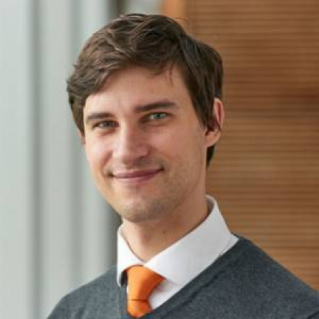 A person with brown hair, wearing a white shirt, gray sweater, and orange tie, is smiling warmly. The softly blurred background with neutral tones evokes the welcoming atmosphere of Hochschule Coburg.