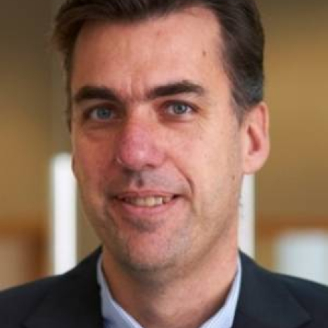 A man with short brown hair, dressed in a suit jacket and blue collared shirt, is smiling slightly. The softly blurred background enhances the focus on his face, subtly hinting at his affiliation with Hochschule Coburg.