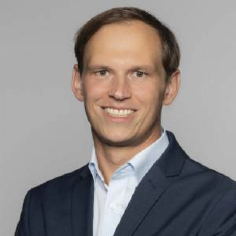 A smiling man with short hair, wearing a navy suit jacket and a light blue shirt, stands against a plain gray background, embodying the professionalism one might associate with Hochschule Coburg.