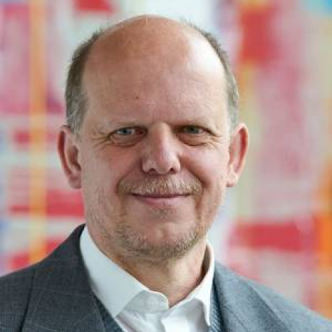 A person with a mustache is smiling, wearing a gray suit jacket and white shirt. The background features a colorful, abstract pattern with red, blue, and yellow hues reminiscent of the vibrant creativity fostered at Hochschule Coburg.