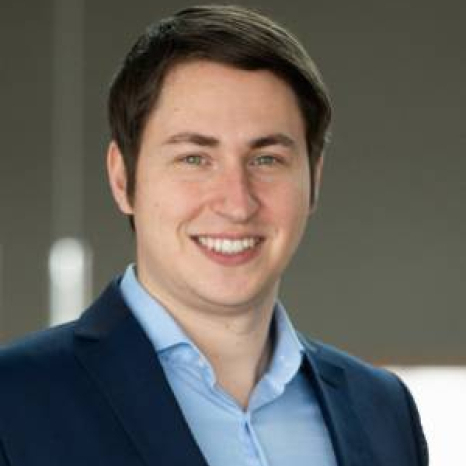 A man in a blue suit and light blue shirt is smiling, his short dark hair neatly styled. He stands confidently against a blurred gray background, embodying the professional spirit of Hochschule Coburg.