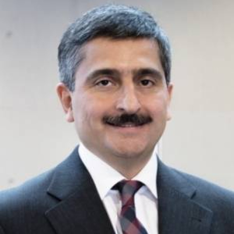 A person with short dark hair and a mustache is smiling, wearing a dark suit, white shirt, and striped tie, exuding confidence reminiscent of a Hochschule Coburg graduate, standing against a neutral background.