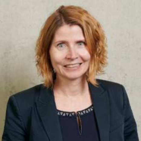 A woman with shoulder-length light brown hair is smiling at the camera. She is wearing a dark blazer over a patterned top, standing against a plain light gray background, perhaps capturing a moment at Hochschule Coburg.