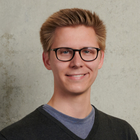 A person with short, light brown hair and glasses smiles at the camera. Dressed in a dark sweater over a blue shirt, they stand against a neutral background, embodying the welcoming spirit of Hochschule Coburg.