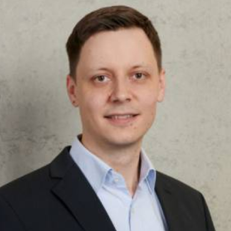 A man in a dark suit and light blue shirt smiles slightly against a plain light gray wall. Representing Hochschule Coburg with confidence, he has short brown hair and is looking directly at the camera.