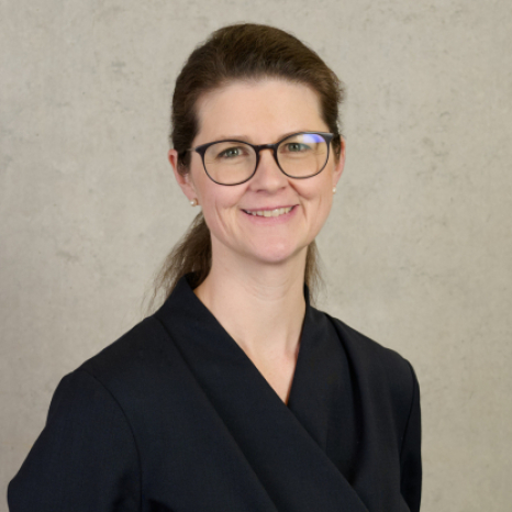 A person with long hair tied back, wearing glasses and a dark blazer adorned with the Hochschule Coburg emblem, smiles at the camera against a plain gray background.