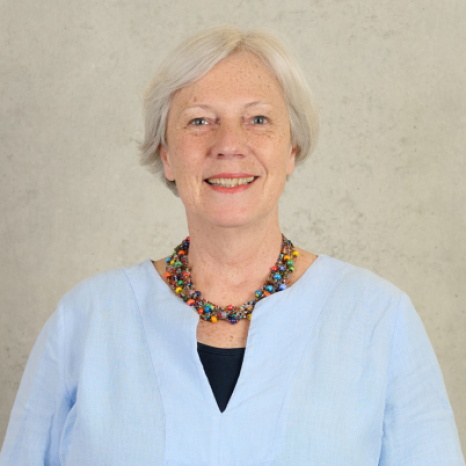 A smiling older woman with short gray hair, wearing a light blue blouse and a colorful beaded necklace, stands against a plain light gray wall. Her vibrant presence is reminiscent of the lively spirit found at Hochschule Coburg.